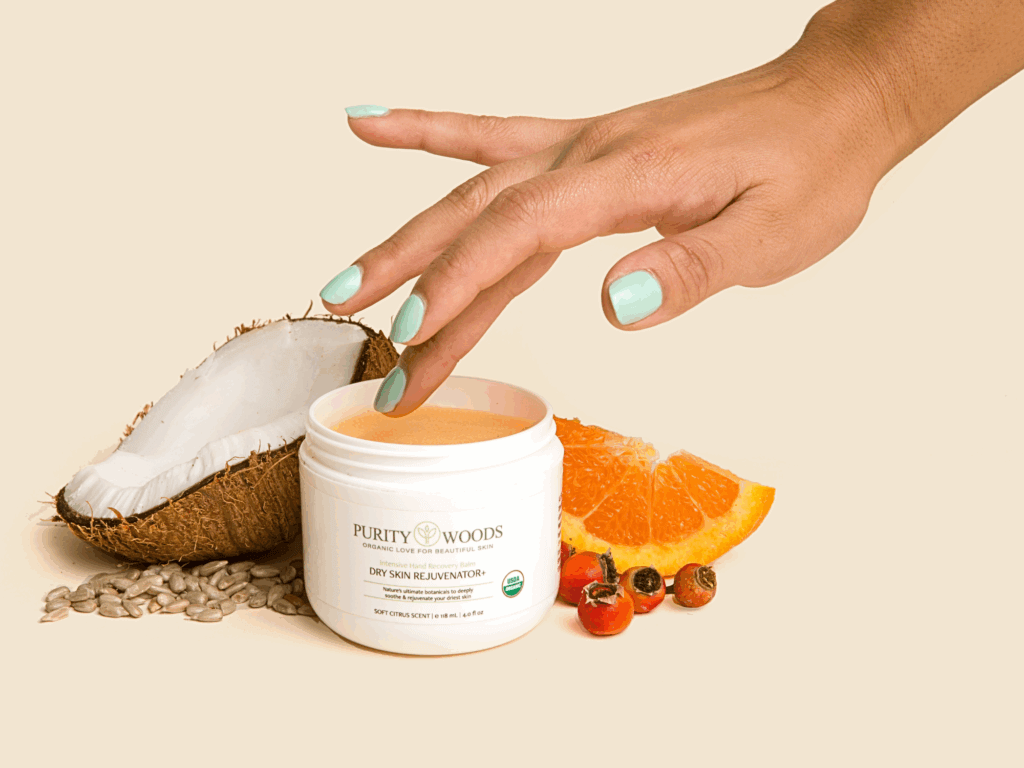 Hand with mint green nails touching peach-colored cream in open ‘Purity Woods Dry Skin Rejuvenator’ jar, surrounded by natural ingredients like coconut, orange slice, sunflower seeds, and rose hips.