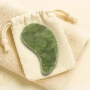 A green jade gua sha facial massage tool rests atop a small drawstring pouch, placed on a soft beige towel.