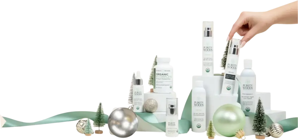 A hand reaches to pick up a bottle labeled "Purity Woods Age-Defying Night Cream" from a festive display of skincare and supplement products. The items are arranged on white cylindrical platforms and include creams, cleansers, exfoliators, and supplements, all branded "Purity Woods" with organic certification. The scene is decorated with miniature Christmas trees, metallic ornaments, and a green ribbon, evoking a holiday theme.