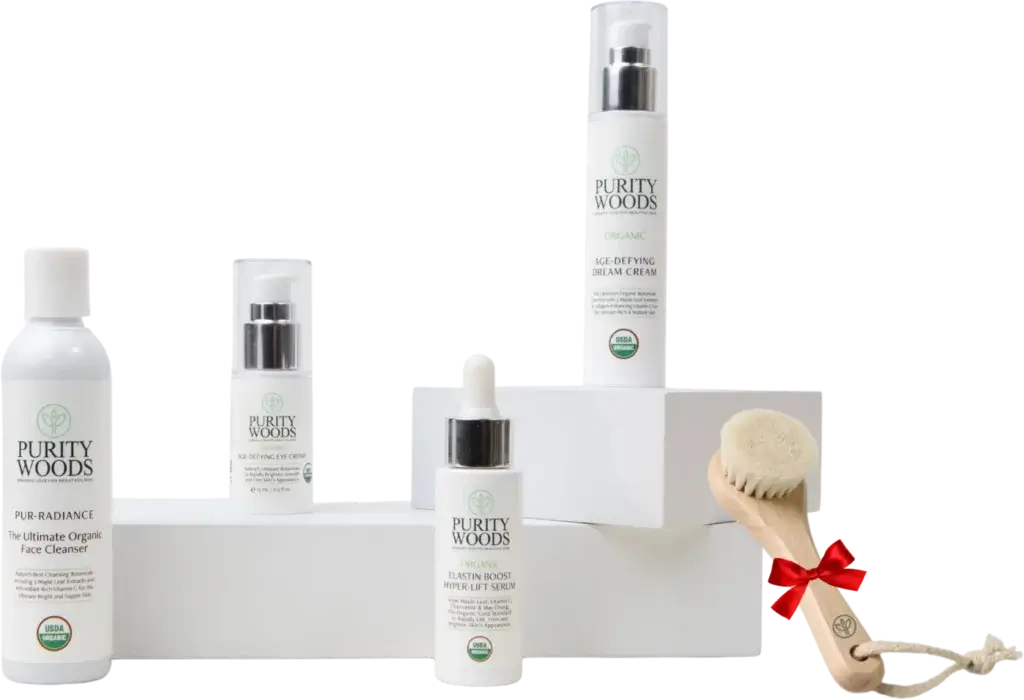 A collection of Purity Woods skincare products arranged on white blocks against a dark gradient background. The products include an organic face cleanser labeled 'PUR-RADIANCE,' an age-defying eye cream, an organic age-defying dream cream, and an organic elastin boost hyper-lift serum. Each product displays the USDA Organic seal. A wooden facial brush with soft bristles and a red ribbon tied around the handle is also shown, emphasizing a premium, organic, anti-aging skincare line.
